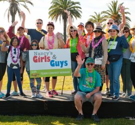 A group of people in leis stand on a platform outdoors, smiling and waving beside a sign reading "Team Contest Winner.