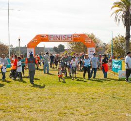 Participants walking under an orange finish line arch at an outdoor event on a sunny day.