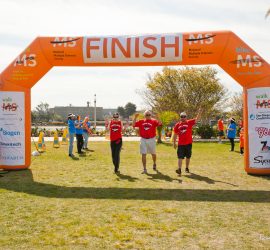 Three people in red shirts cross the "Finish" line at an outdoor MS charity walk event under an orange inflatable arch.