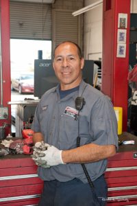 A mechanic in uniform stands in a garage, wearing gloves and holding dirty hands, with tools and equipment in the background.