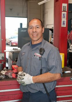 A mechanic in uniform stands in a garage, wearing gloves and holding dirty hands, with tools and equipment in the background.
