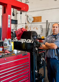 A mechanic stands with folded arms next to a red Snap-on tool chest in an auto repair shop.